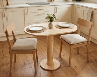 Op de afbeelding: Een ovale eettafel van licht hout met een centrale kolomvoet en twee bijpassende stoelen. Elke stoel is voorzien van een beige stoffen zitkussen dat met touwtjes is vastgemaakt. De tafel is gedekt met twee witte borden op gevlochten placemats.