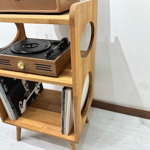 May include: A wooden record player stand with three shelves and two circular cutouts. The stand is brown and has a retro design. A record player and vinyl records are on the shelves.