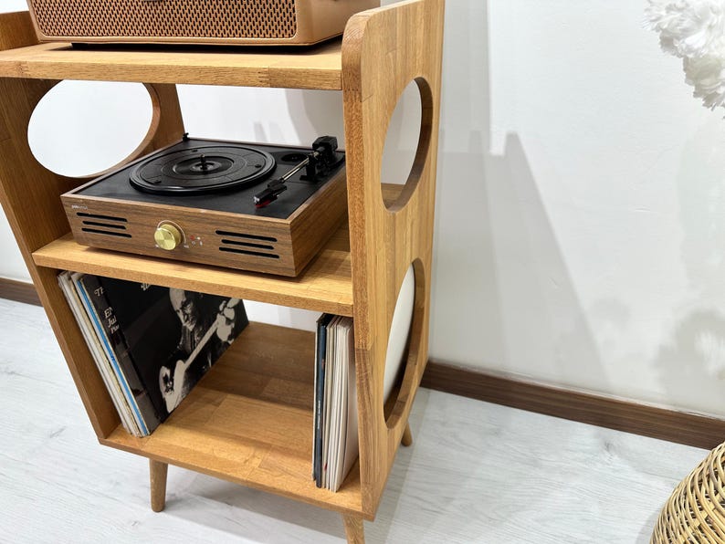 May include: A wooden record player stand with three shelves and two circular cutouts. The stand is brown and has a retro design. A record player and vinyl records are on the shelves.