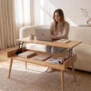 May include: A light-toned wooden coffee table featuring a lift-top mechanism that elevates the surface. The table has four tapered legs and an open storage compartment underneath the main tabletop, which currently holds books and a magazine.