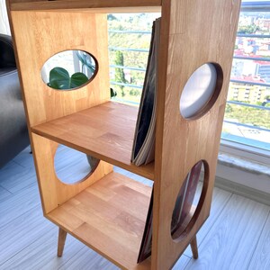 May include: A light-toned wooden storage unit with three tiers, featuring oval cutouts on the side panels. The unit stands on four tapered legs and holds vinyl records on the middle and bottom shelves, with a record player on the top surface.