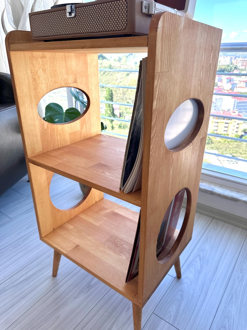 May include: A light-toned wooden storage unit with three tiers, featuring oval cutouts on the side panels. The unit stands on four tapered legs and holds vinyl records on the middle and bottom shelves, with a record player on the top surface.