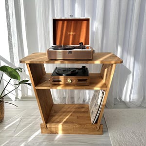 Könnte beinhalten: Ein Holz-Plattenspieler-Ständer mit zwei Plattenspielern. Der obere Plattenspieler befindet sich in einem Koffer im Vintage-Stil. Der Ständer hat zwei Regale und ist aus hellem Holz gefertigt. Links ist eine Topfpflanze.
