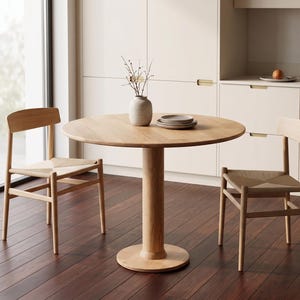 May include: A round wooden dining table with a single pedestal base, accompanied by two wooden chairs with woven seats. A vase with branches and a stack of plates are on the table. The chairs and table are made of light-colored wood.