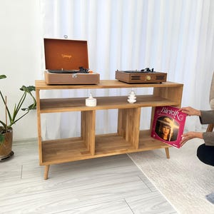 Puede incluir: Mueble de madera para tocadiscos con dos tocadiscos y un disco de vinilo. El mueble tiene tres compartimentos abiertos y patas anguladas. Un tocadiscos es marrón con un exterior estampado, el otro es de madera más oscura.