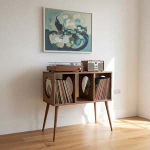 May include: A wooden record storage console with four tapered legs, featuring two rectangular compartments filled with vinyl records and two circular side cutouts. A turntable and a radio are placed on the top surface of the wooden unit.