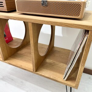 Puede incluir: Un soporte de madera para tocadiscos con cuatro estantes y patas de metal negro. El soporte tiene un recorte redondeado en cada estante. El estante superior tiene un tocadiscos encima. El estante inferior tiene una pila de discos de vinilo.