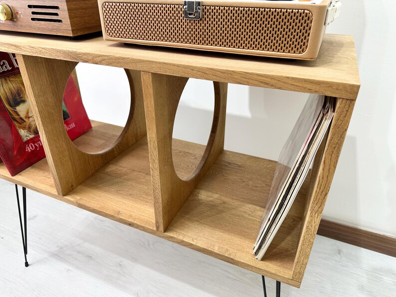 Puede incluir: Un soporte de madera para tocadiscos con cuatro estantes y patas de metal negro. El soporte tiene un recorte redondeado en cada estante. El estante superior tiene un tocadiscos encima. El estante inferior tiene una pila de discos de vinilo.