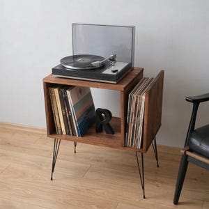 May include: A wooden record player stand with a black turntable on top. The stand has two compartments for vinyl records and sits on black hairpin legs. A black letter "R" is on the shelf.