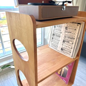 May include: A light brown wooden record player stand with three shelves and circular cutouts on the side panels. A beige portable record player with the word Vintage printed on the lid sits on the top shelf, while vinyl records are stored below.