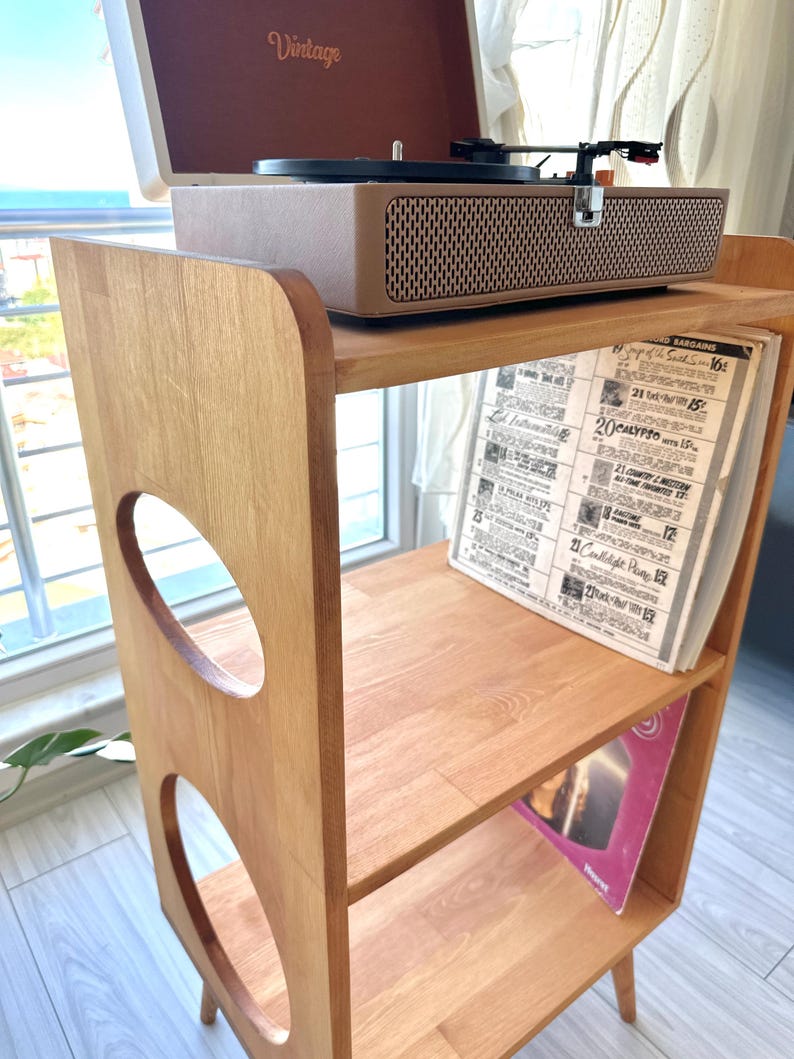 May include: A light brown wooden record player stand with three shelves and circular cutouts on the side panels. A beige portable record player with the word Vintage printed on the lid sits on the top shelf, while vinyl records are stored below.