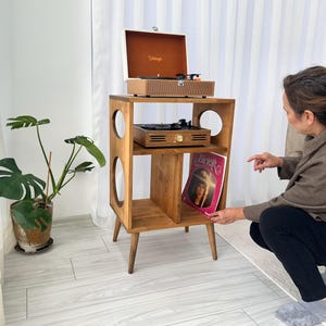 May include: A wooden record player stand with a vintage record player on top and a record album on the shelf. The stand has a mid-century modern design with circular cutouts and tapered legs. A potted plant is to the left.