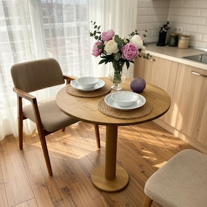 Puede incluir: Una pequeña mesa de comedor redonda de madera con dos sillas. La mesa está puesta con dos cuencos blancos sobre manteles individuales tejidos. Un jarrón de peonías rosas y blancas está en el centro. Las sillas tienen asientos de tela beige y marcos de madera.