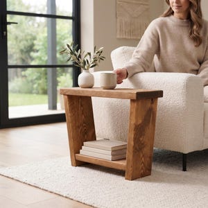 May include: A rectangular wooden side table with a dark brown finish, featuring angled side supports and a lower shelf holding a stack of three books. The top surface holds a small white ceramic vase with green leaves and a beige ceramic mug.