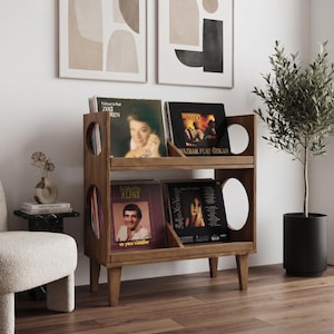 Può includere: Uno scaffale in legno a due ripiani per dischi in vinile, caratterizzato da aperture laterali circolari e quattro gambe affusolate. L'unità contiene dischi con copertine visibili, tra cui Zeki Müren, Mazhar Fuat Özkan, Alpay e una custodia nera.