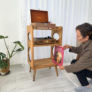 Może przedstawiać: Drewniany stojak na gramofon z dwoma gramofonami i walizką na gramofon w stylu vintage. Stojak ma konstrukcję z trzema półkami z okrągłymi wycięciami. Osoba trzyma płytę z napisem "Bicicik".
