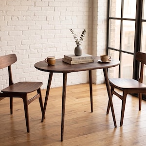 Pode incluir: Uma mesa de jantar oval de madeira escura com quatro pernas cónicas e duas cadeiras de madeira a condizer. Sobre a mesa estão duas canecas de cerâmica, dois livros intitulados The Loft e Mid-Century Forms e um pequeno vaso cinzento.