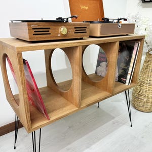 Puede incluir: Un soporte de madera para tocadiscos con tres estantes y patas de horquilla negras. El soporte tiene dos recortes circulares en el estante superior y contiene un tocadiscos, un altavoz y discos de vinilo.