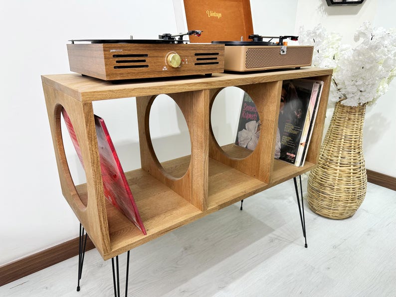 Puede incluir: Un soporte de madera para tocadiscos con tres estantes y patas de horquilla negras. El soporte tiene dos recortes circulares en el estante superior y contiene un tocadiscos, un altavoz y discos de vinilo.