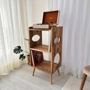 May include: A wooden record player stand with a vintage-style record player on top. The stand has two shelves with circular cutouts and angled legs. A vinyl record is visible on the lower shelf. The stand is made of light-colored wood.