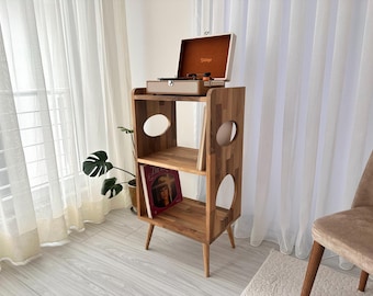Könnte beinhalten: Ein hölzerner Plattenspielerständer mit einem Plattenspieler im Vintage-Stil obenauf. Der Ständer hat zwei Regale mit kreisförmigen Aussparungen und abgewinkelten Beinen. Eine Schallplatte ist auf dem unteren Regal sichtbar. Der Ständer ist aus hellem Holz gefertigt.
