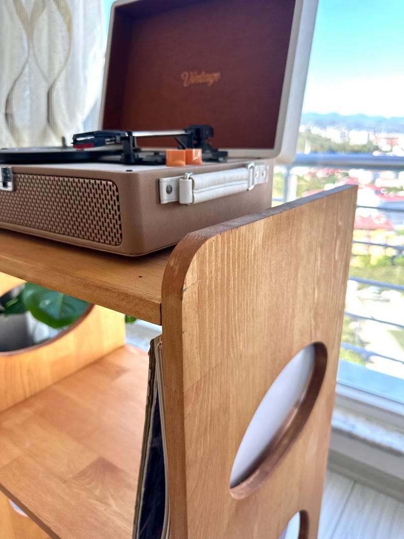 May include: A light brown wooden record player stand with oval cutouts on the side panels and a shelf for vinyl records. A beige and brown portable record player with the word Vintage printed on the inside lid sits on the top surface.