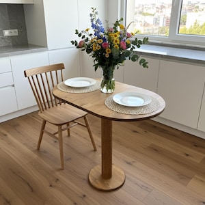 Puede incluir: Pequeña mesa ovalada de madera con una base de pedestal, preparada para dos con platos blancos y manteles individuales tejidos. Una silla de madera está al lado de la mesa. Un jarrón con flores coloridas está sobre la mesa.