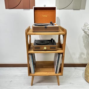May include: A wooden record player stand with three shelves. The stand has a vintage record player on the top shelf, a turntable on the middle shelf, and vinyl records on the bottom shelf. The stand has a light wood finish and rounded edges.