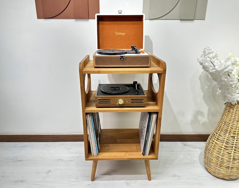May include: A wooden record player stand with three shelves. The stand has a vintage record player on the top shelf, a turntable on the middle shelf, and vinyl records on the bottom shelf. The stand has a light wood finish and rounded edges.