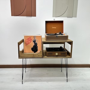 May include: A wooden record player stand with thin black metal hairpin legs, featuring a two-tier design. The top shelf holds an open brown vintage-style turntable, while the lower section displays a vinyl record cover featuring Emel Sayin.