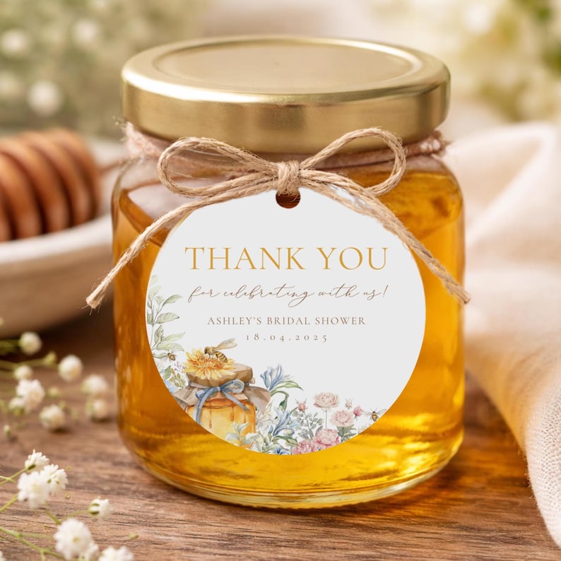 May include: A small glass jar filled with golden honey, topped with a gold lid and a decorative tag. The tag reads "THANK YOU" and includes floral illustrations and event details. Tied with twine, the jar is a charming gift.