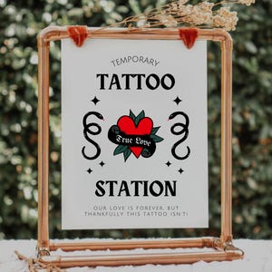 K&ouml;nnte beinhalten: Ein wei&szlig;es Schild mit schwarzem Text und einem roten Herz-Tattoo-Design lautet "Tempor&auml;re Tattoo-Station. Unsere Liebe ist f&uuml;r immer, aber zum Gl&uuml;ck ist dieses Tattoo nicht so."