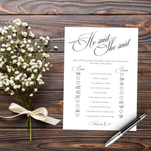 Puede incluir: Una tarjeta de juego rectangular blanca titulada He said She said con texto negro e iconos de coraz&oacute;n, acompa&ntilde;ada de un ramo de flores de paniculata blancas atadas con una cinta crema y un bol&iacute;grafo negro y plateado sobre madera.