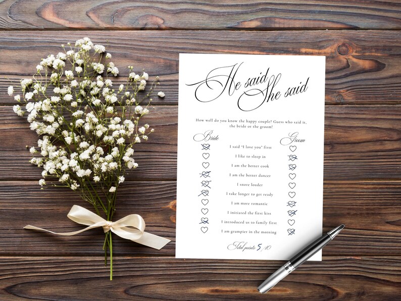 Puede incluir: Una tarjeta de juego rectangular blanca titulada He said She said con texto negro e iconos de coraz&oacute;n, acompa&ntilde;ada de un ramo de flores de paniculata blancas atadas con una cinta crema y un bol&iacute;grafo negro y plateado sobre madera.