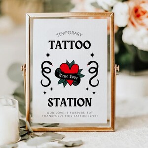 K&ouml;nnte beinhalten: Ein goldfarbener Rahmen mit dem Text "Temporary Tattoo Station" und einem roten Herzen mit den Worten "True Love" umgeben von Schlangen. Das Schild ist auf wei&szlig;em Hintergrund mit schwarzen Sternen.