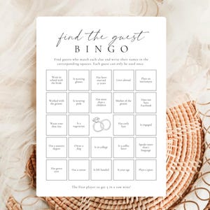 Puede incluir: Una tarjeta de juego de bingo Find the Guest rectangular blanca con texto negro y una ilustración central de un anillo de diamantes. La cuadrícula contiene veinticinco cuadrados con varias pistas sobre los invitados, instrucciones y una regla.