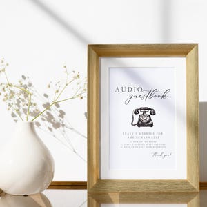 K&ouml;nnte beinhalten: Ein gerahmtes Schild mit den Worten "Audio Guestbook" in eleganter Schrift, mit einer Vintage-Telefon-Illustration. Das Schild enth&auml;lt Anweisungen zum Hinterlassen einer Nachricht f&uuml;r das Brautpaar. Der Rahmen ist hellgoldfarben.