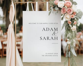 Peut inclure: Un panneau de bienvenue rectangulaire blanc suspendu à un cadre en métal doré. Le panneau porte le texte noir WELCOME TO OUR HAPPILY EVER AFTER, ADAM & SARAH, 12 AUGUST 2024 et THE ORCHARD ESTATE. Un arrangement floral de roses roses et de feuillage orne le coin supérieur droit.