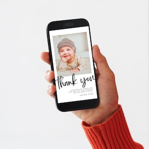 May include: A smartphone screen shows a baby photo with the words "Thank you" in black script. The phone is held in a hand wearing an orange jumper. The baby wears a striped hat and smiles.