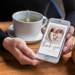 May include: A smartphone displays a baby announcement with two newborns in white outfits. The screen reads "Thank you" in elegant script. A white teacup with tea and a saucer are in the background.