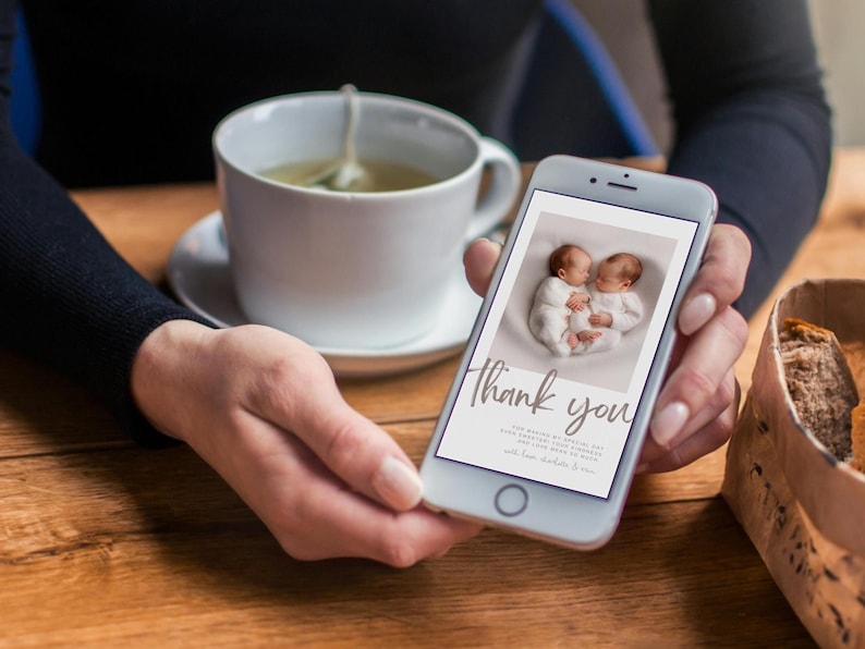 May include: A smartphone displays a baby announcement with two newborns in white outfits. The screen reads "Thank you" in elegant script. A white teacup with tea and a saucer are in the background.
