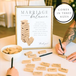 Puede incluir: Un letrero enmarcado en dorado con el texto "MATRIMONIO es un juego de equilibrio" e instrucciones para los invitados. Se muestra un juego tipo Jenga con bloques de madera. El letrero incluye un corazón hecho de bloques. El letrero dice "VIENE EN TRES TAMAÑOS".