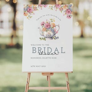 Op de afbeelding: Een wit bord op een ezel met bloemversieringen en de tekst "Welkom bij de Bridal Shower" en "Ter ere van Juliette Rose". Een theepotillustratie is gecentreerd boven de tekst. De datum "16 mei 2025" staat onderaan.