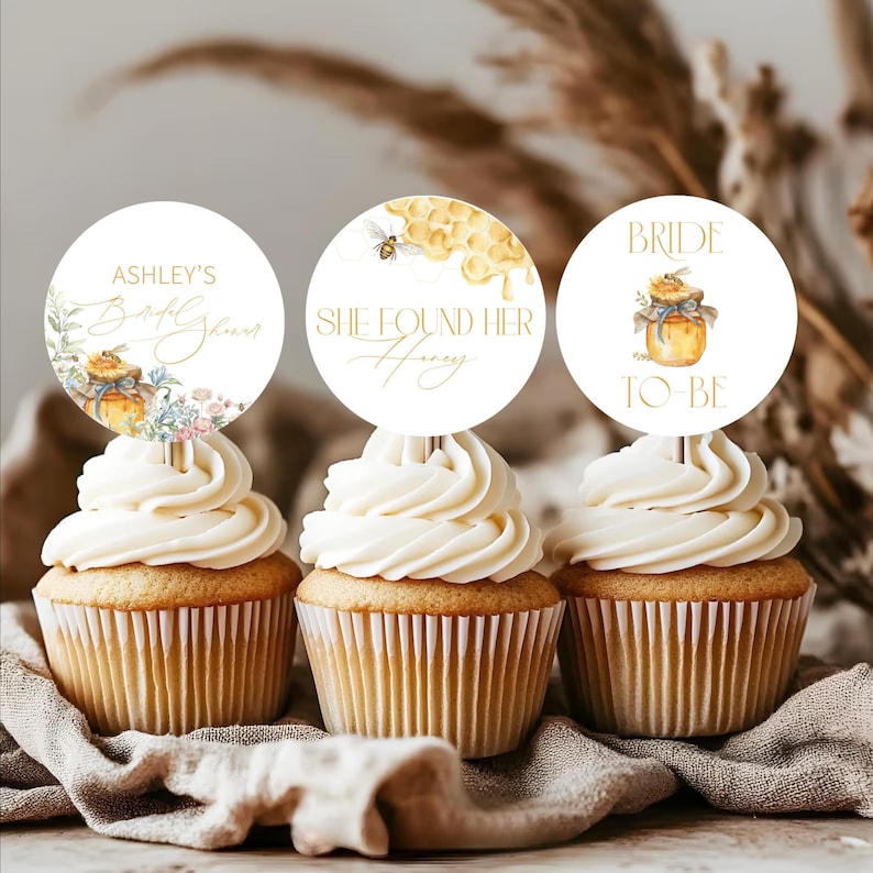 May include: Three cupcakes with white frosting and decorative toppers. The toppers have text and illustrations related to a bridal shower theme. One says "Ashley's Bridal Shower", another "She Found Her Honey", and the last "Bride To Be".