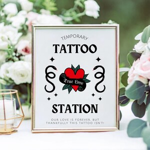 K&ouml;nnte beinhalten: Ein wei&szlig;es Schild mit schwarzem Text, auf dem "Temporary Tattoo Station" steht, mit einem roten Herzen und schwarzen Schlangen darauf. Das Schild ist in Gold gerahmt und steht auf einem Tisch mit Blumen.