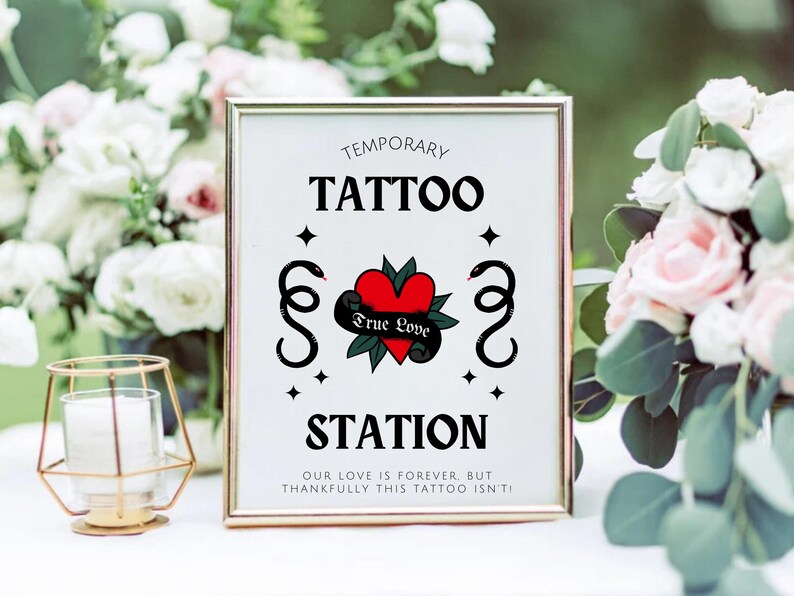 K&ouml;nnte beinhalten: Ein wei&szlig;es Schild mit schwarzem Text, auf dem "Temporary Tattoo Station" steht, mit einem roten Herzen und schwarzen Schlangen darauf. Das Schild ist in Gold gerahmt und steht auf einem Tisch mit Blumen.