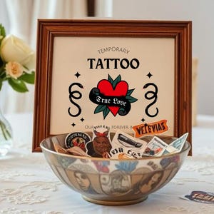 K&ouml;nnte beinhalten: Ein gerahmtes Schild mit dem Text "Temporary Tattoo" und einem Herzen mit den Worten "True Love" darin. Das Schild steht auf einem Tisch mit einer Sch&uuml;ssel voller tempor&auml;rer Tattoos.