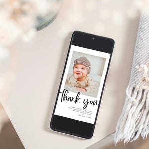 May include: A smartphone displays a photo of a smiling baby wearing a striped hat. The screen features the words "Thank you" in elegant script, with additional text below. The phone rests on a white surface, next to a gray blanket and dried flowers.