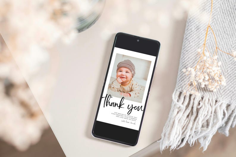 May include: A smartphone displays a photo of a smiling baby wearing a striped hat. The screen features the words "Thank you" in elegant script, with additional text below. The phone rests on a white surface, next to a gray blanket and dried flowers.
