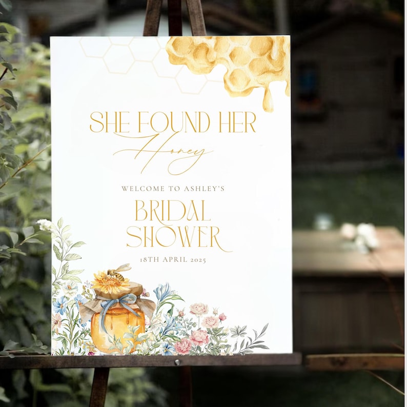 May include: A white sign with gold text that reads "She Found Her Honey" and "Welcome to Ashley's Bridal Shower". The sign has a watercolor illustration of a honey pot and flowers. The date "18th April 2025" is also displayed.